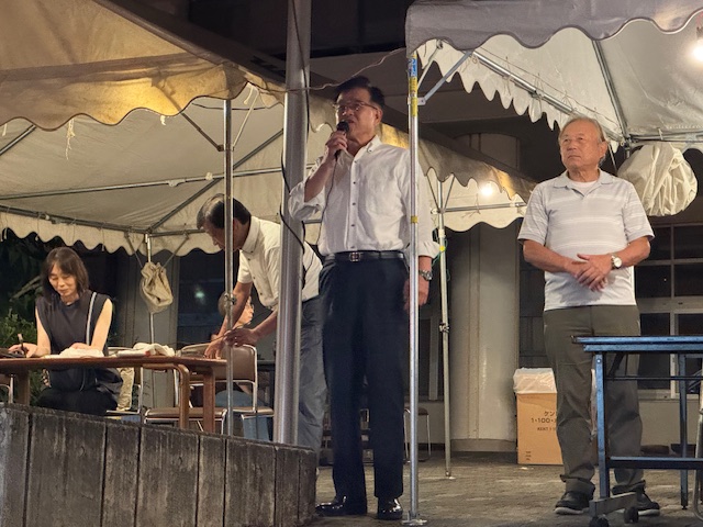 夜の屋外会場のテント内で、白いシャツ姿の市長がマイクを持ってスピーチしており、横には年配の男性が立っており、手前には座っている女性が見える納涼大会の写真