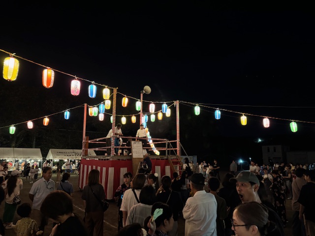 提灯が飾られたやぐらの上で、祭りの関係者と思われる人々が立っており、その周囲を多くの浴衣や私服姿の住民が囲んで夜の盆踊りを楽しんでいる様子の写真