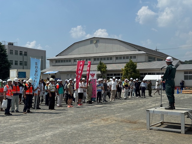 屋外の校庭で、ヘルメット姿の市長が演台の上でマイクを持ち、防災訓練に参加者に訓示を述べている様子の写真