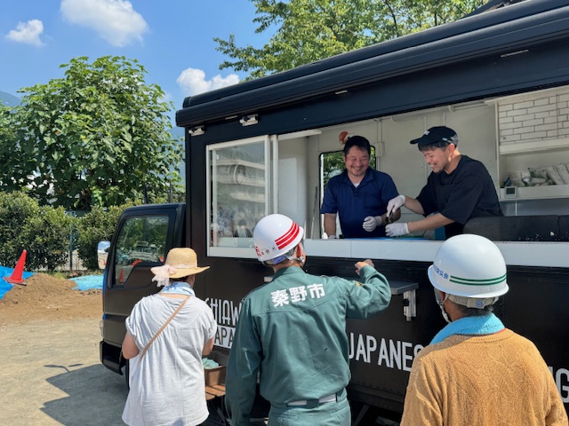 黒いキッチンカーの窓口で、笑顔の店員から防災訓練の参加者と思われるヘルメット姿の市長と一般の高齢者が炊き出しの食べ物を受け取っている様子の写真