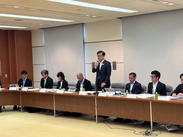 会議室の長机に座った複数のスーツ姿の男女の前で、中央のスーツ姿の市長がマイクを持ち、身振り手振りを交えながら熱心にスピーチをしている様子の写真