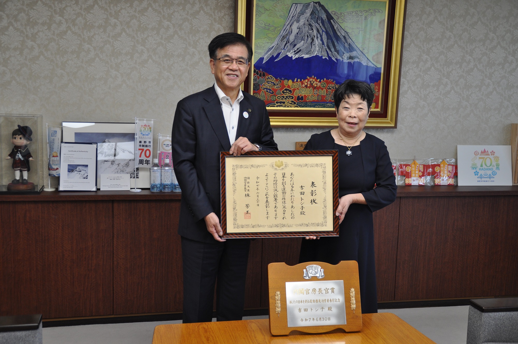 壁に富士山の絵画が飾られた室内で、スーツ姿の市長と黒いワンピース姿の吉田トシ子さんが、「内閣官房長官表彰」と書かれた盾の置かれたテーブルの奥に立ち、賞状の額縁を一緒に持っている記念写真