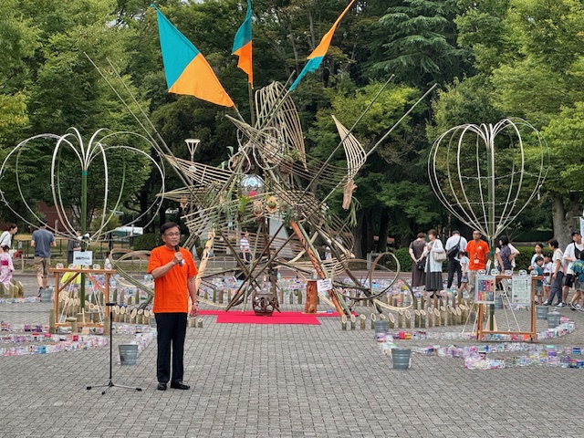 公園の広場の竹や金属のフレームなどで作られた抽象的なオブジェの前で、オレンジ色のTシャツを着た市長がマイクを持ち、大勢の聴衆に向かってスピーチをしているイベントの様子の写真