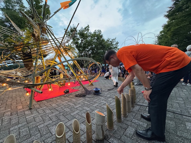 公園の広場で開催されているイベントで、オレンジ色のTシャツを着た市長が竹筒にろうそくを立てて火を灯す準備をしている夕方の様子の写真