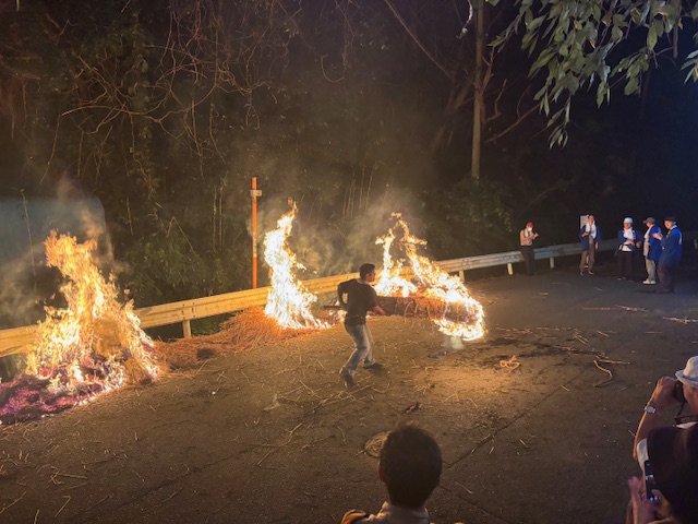 山道の夜間で、舗装された路上で激しく燃え上がる炎の間を、一人の男性が燃える藁束のようなものを持って走る、勇敢な火祭りの様子の写真