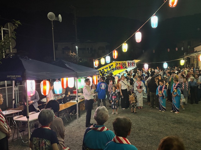 夜間の広場で行われている祭り会場で、提灯が飾られたテントの前で白いシャツ姿の市長がマイクを持ち、浴衣姿の子どもや大人など大勢の観客に向かって挨拶をしている様子の写真