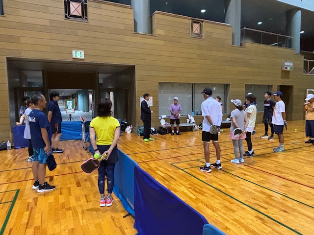 体育館の木製フロアで、スーツ姿の市長の周りに、ラケットを持った数名の参加者が立っている練習会の様子の写真