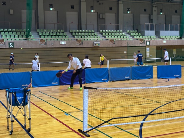 体育館の観客席が奥に見える広いフロアで、複数の参加者たちが數面のコートに分かれてピックルボールをしている手前のコートで白いシャツ姿の市長がピックルボールを練習している写真