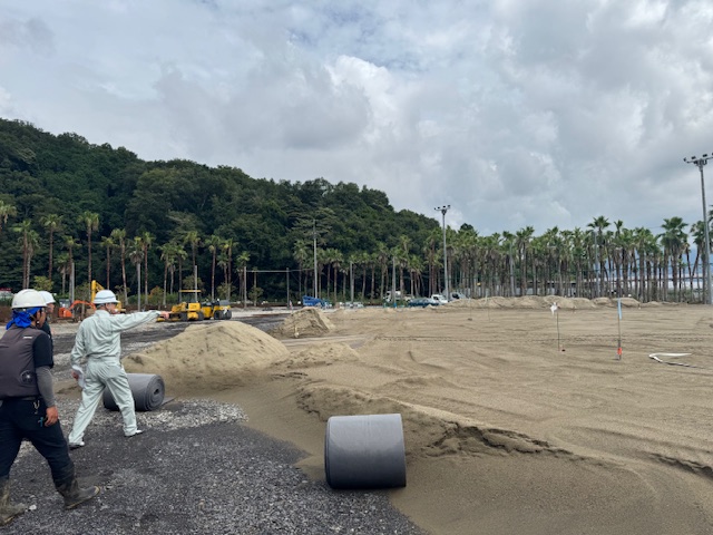 曇り空の下、ヤシの木が並ぶ背景で、砂利と大量の砂が敷かれた広大な敷地に、作業服姿の男性2人が立ち、一人が指を差しながら現場の指示をしている建設工事の様子の写真