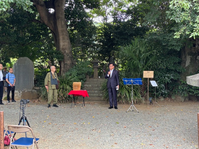 木々が生い茂る屋外の砂利の敷かれた会場で、スーツ姿の市長がマイクを持ち、左側には作務衣のような服装の年配の男性が立っている、供養会の様子の写真