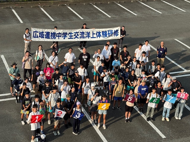 「広域連携中学生交流洋上体験研修」と書かれた横断幕を掲げた大人たちと、手前で数字のプラカードを持つ多数の中学生が駐車場で集合している俯瞰の記念写真