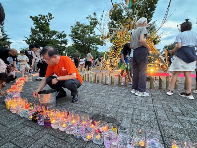 夕暮れの公園の広場で、オレンジ色のTシャツを着た市長が地面に並べられたペットボトルを再利用したグラスに入ったろうそくに明かりを灯している、イベントの様子の写真