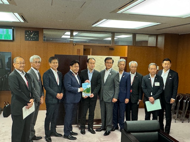 木目調の壁の会議室で、市長を含むスーツ姿の男性10名が横一列に並んでおり、中央の男性が資料と思われる冊子を掲げてカメラに向かって立っている集合写真