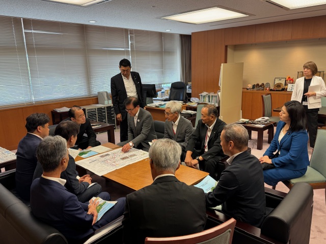 木目調の応接室の大きなローテーブルを囲み、スーツ姿の男女が着席または起立し、市長が地図のような大きな資料を広げて議論している会議の様子の写真