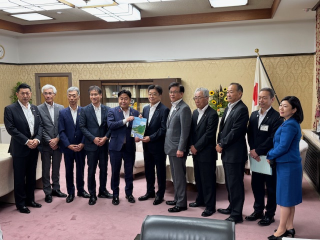 日本の国旗が立てられた木目調の応接室で、市長を含むスーツ姿の男女12名が横一列に並んでおり、中央の男性が資料と思われる冊子を掲げて立っている集合写真