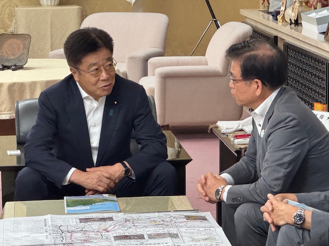 応接室のソファに座ったスーツ姿の市長と議員バッチをつけた男性が、広げられた地図のような資料を挟んで顔を寄せ合い、真剣な表情で話し合っている様子の写真