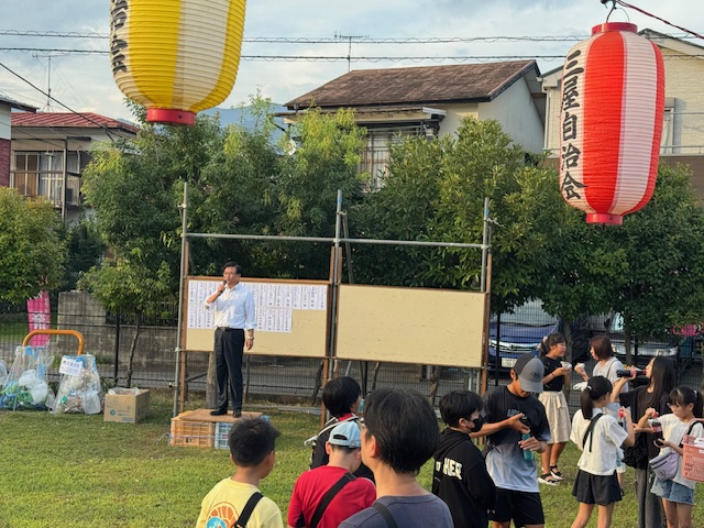 住宅地の中の広場で、白いシャツ姿の市長が足場の上でマイクを持ち、黄色と赤の提灯が飾られた前で、集まった大勢の子どもや住民に向かって挨拶をしている祭りの様子の写真