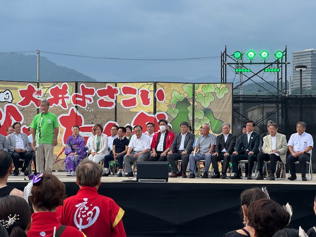 外の特設ステージで、緑色のポロシャツを着た男性がマイクを持ちスピーチをしており、その後ろには浴衣姿の女性やスーツ姿の男性を含む多くの来賓が着席している祭りの様子の写真