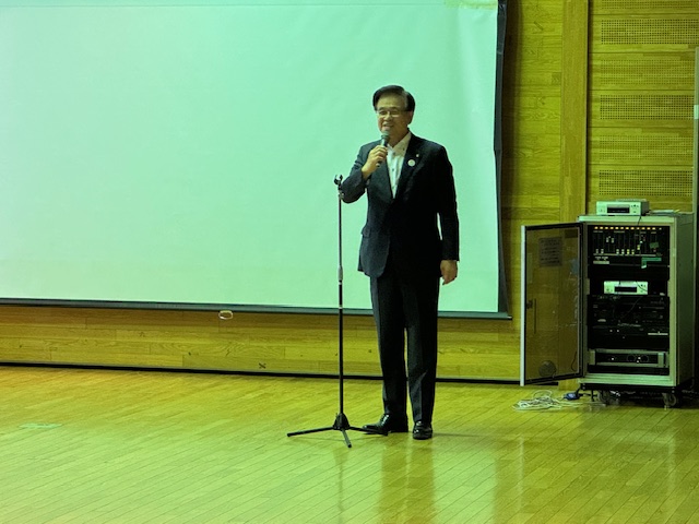 木製の床の会場のステージ上で、スーツ姿の市長がマイクを持って立ち、背後の大きなスクリーンを前にスピーチをしている社交ダンス大会の様子の写真