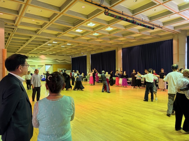 明るい照明の広いホールで、多くの参加者が思い思いの衣装を身につけ、社交ダンスを楽しんでいる様子を、手前の市長と女性が見つめているダンス大会の様子の写真