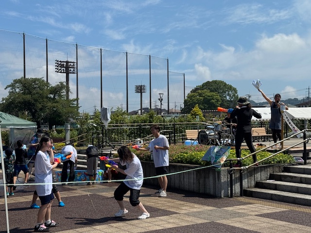 青空の下、広場のステージと階段がある場所で、若者たちが水鉄砲を撃ち合って遊んでおり、右奥のステージ上にはタオルを振る人が立っている水かけイベントの様子の写真