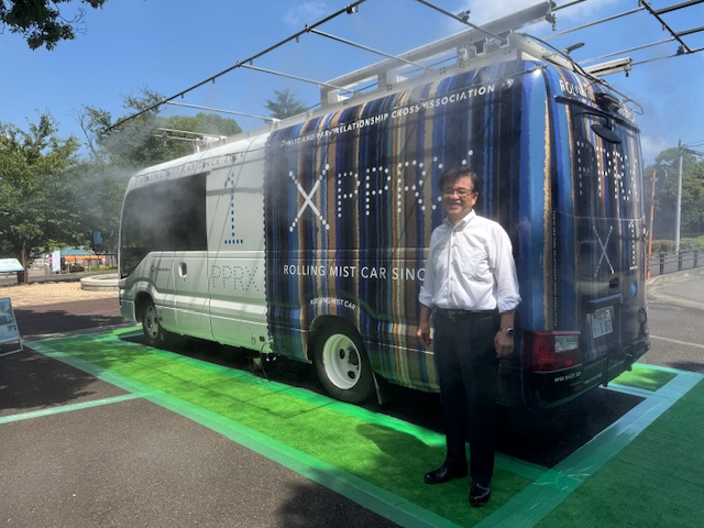 白と青のグラフィックで装飾された「ROLLING MIST CAR SINCE 2017」と書かれたミストカーの前で、白いシャツと黒いスラックス姿の市長が笑顔でカメラに向かって立っている写真