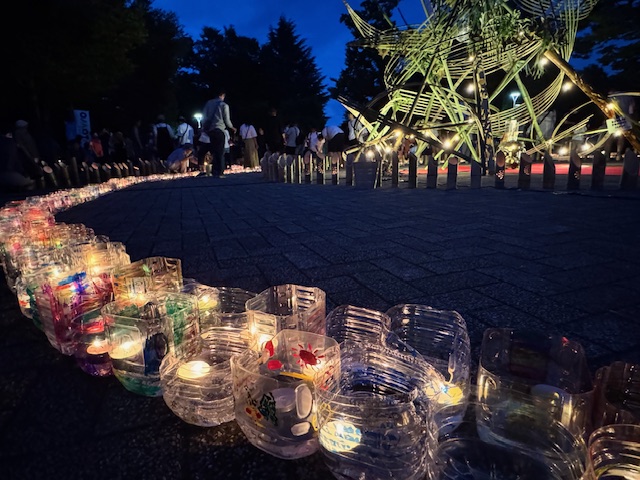 夜の公園の広場で、ペットボトルを再利用したグラスに入れたろうそくが長く連なって道を照らしており、奥には竹と電飾で作られた大きなオブジェが見える幻想的なイベントの様子の写真