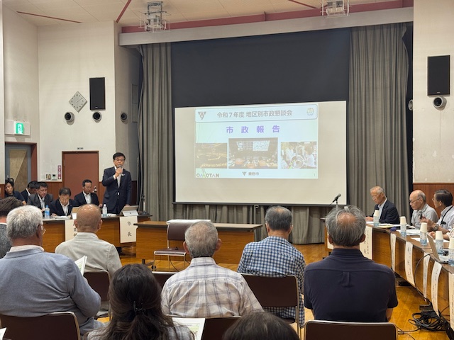 「令和7年度 地区別市政懇談会 市政報告」の資料が投影されたスクリーンを背に、スーツ姿の市長が、大勢の高齢者を含む市民や関係者の前でマイクを持って話している懇談会の様子の写真