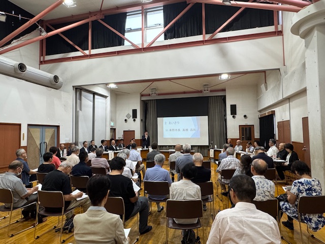 体育館のような広い会場で、スクリーンに「あいさつ 秦野市長 高橋 昌和」と表示された前で、スーツ姿の市長がマイクを持って挨拶をしているところを大勢の参加者が椅子に座っている後方から捉えた写真