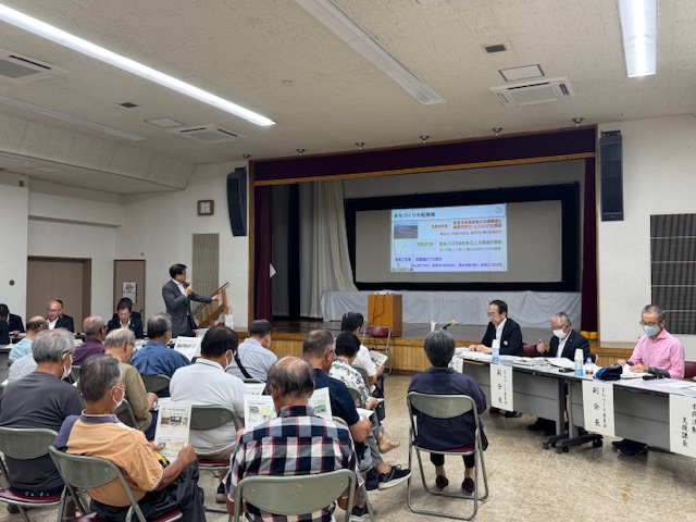 会議室のステージ上で、大勢の高齢者を含む参加者の前にスーツ姿の市長が立ち、スクリーンに資料を投影しながら説明をしている懇談会の写真