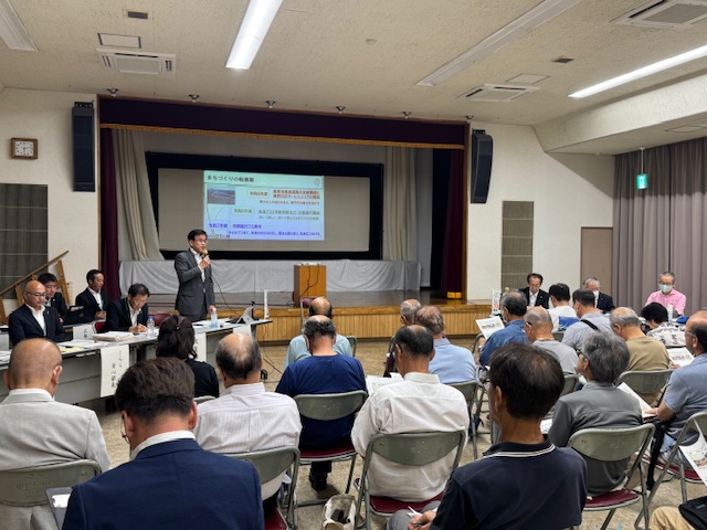 会議室のステージ上で、スーツ姿の市長がスクリーンに資料を投影しながらマイクを持って説明をしており、大勢の高齢者を含む参加者がそれを熱心に聴講している懇談会の写真