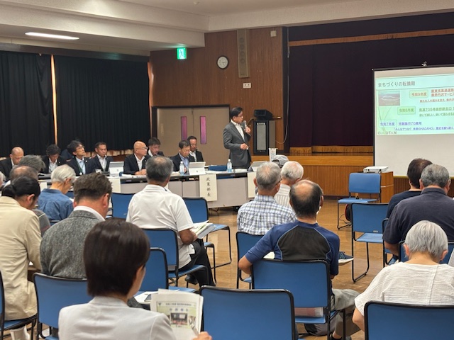 公民館のような会場のステージ上で、大勢の高齢者を含む参加者が着席している前にスーツ姿の市長が立ち、マイクを持ち資料をスクリーンに投影しながら説明をしている懇談会の写真