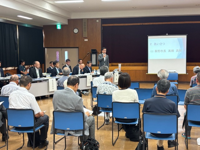 公民館のような会場のステージ上で、スーツ姿の市長がマイクを持って挨拶をしており、大勢の高齢者を含む市民や関係者が着席してそれに耳を傾けている懇談会の様子の写真