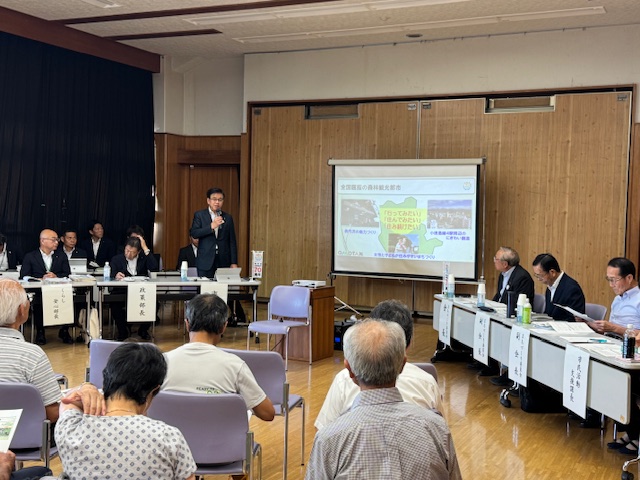 公民館のような会場のステージ上で、大勢の高齢者を含む参加者に向けてスーツ姿の市長がマイクを持ち、スクリーンに資料を投影しながら説明をしている懇談会の写真