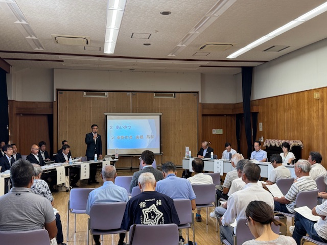 公民館のような会場のステージ上で、大勢の高齢者を含む参加者に向けてスーツ姿の市長がマイクを持ち、挨拶をしている懇談会の写真