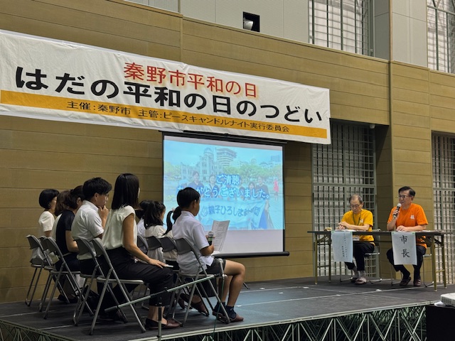 「秦野市平和の日」と書かれた横断幕の下、ステージ上でオレンジ色のTシャツを着た市長がマイクを持ち、左側には数名の参加者がスクリーンを前に着席しているイベントの様子の写真
