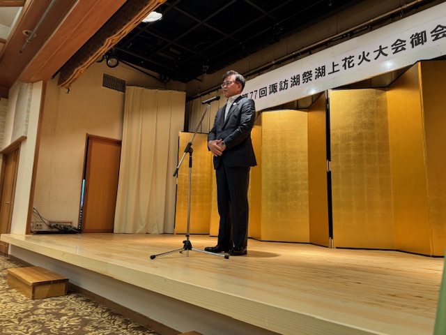 第77回諏訪湖祭湖上花火大会御会」と書かれた立て看板と金屏風のある和風のステージ上で、スーツ姿の市長がマイクスタンドの前でスピーチをしている様子の写真