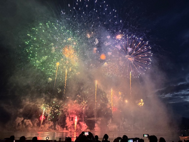 夜の湖上から打ち上げられた色とりどりの花火が空いっぱいに広がり、手前にはその様子を多くの観客がスマートフォンなどで撮影している迫力のある花火大会の様子の写真