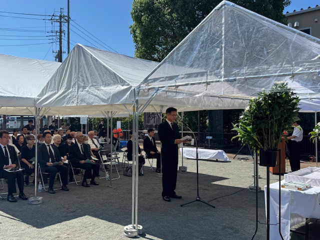 白いテントが張られた屋外で、喪服姿の市長がマイクスタンドの前で弔辞を読み上げており、他の参列者も全員が着席してそれに耳を傾けている平和祈念式典の様子の写真