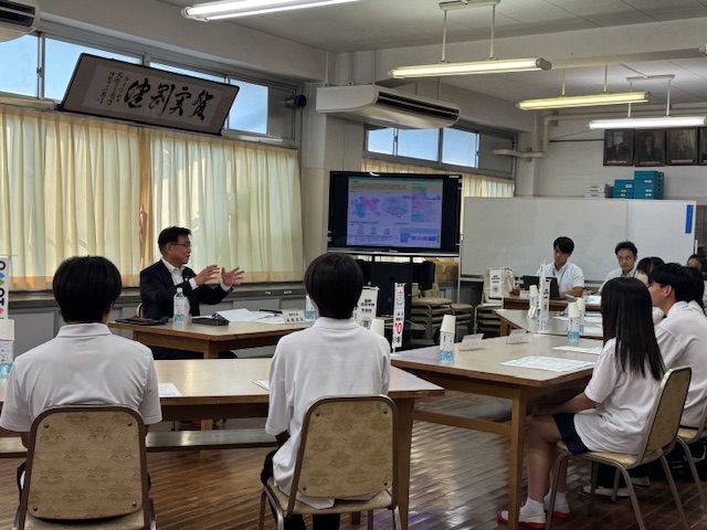 学校の教室で、スーツ姿の市長が数名の引率者と複数の制服姿の高校生を前に、スクリーンに資料を投影しながら何かを熱心に説明している意見交換会の様子の写真