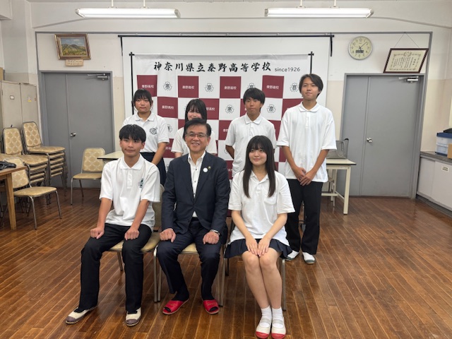 「神奈川県立秦野高等学校 since 1926」と書かれた校名板の前で、スーツ姿の市長が中央に座り、その周りを男子生徒3名と女子生徒3名で囲んで整列している集合写真