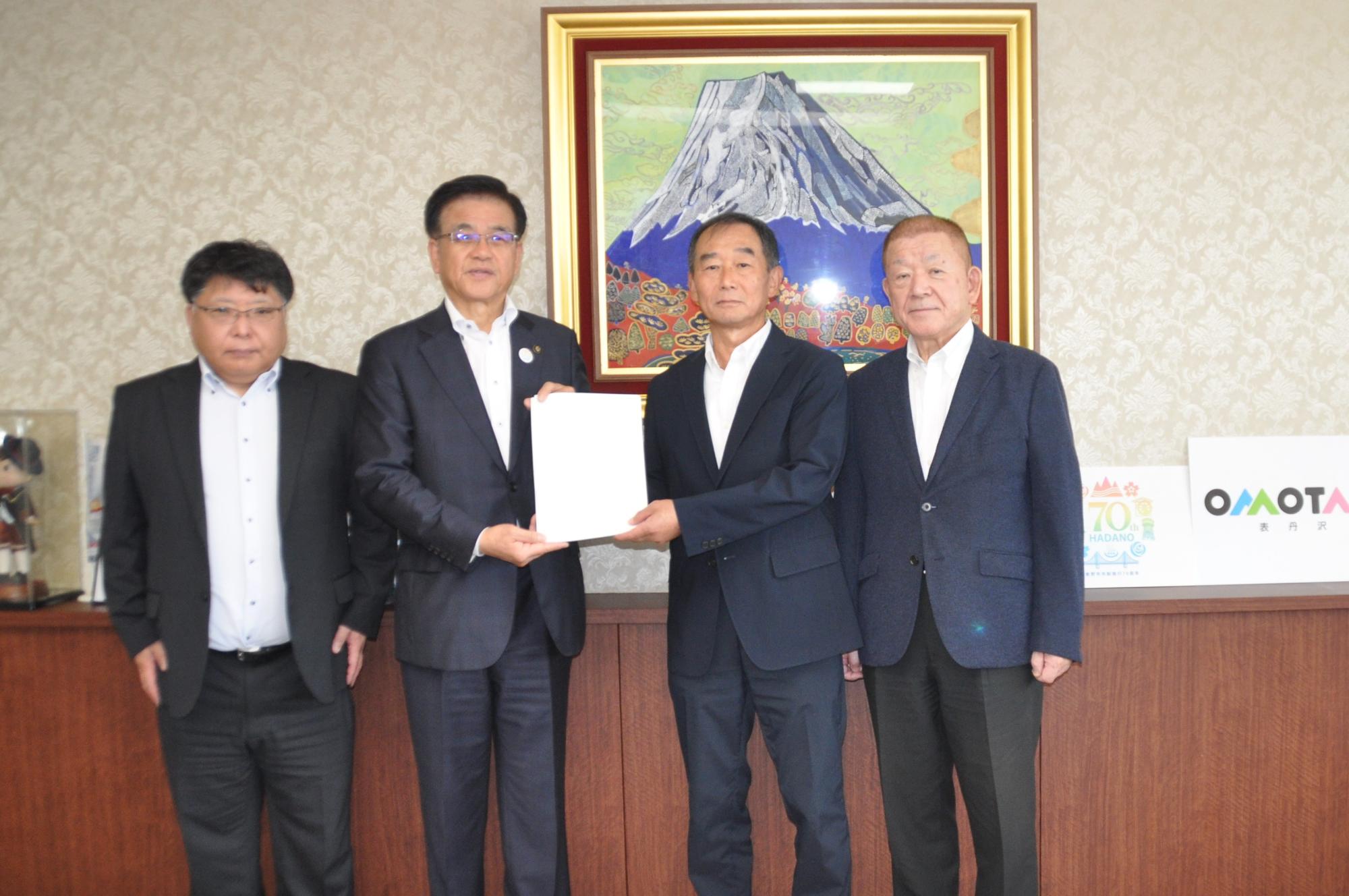 富士山の絵画が飾られた室内で、スーツ姿の4名の男性が並んで立ち、中央の市長と男性が書類を両手で持ちながらカメラに向かって立っている記念写真