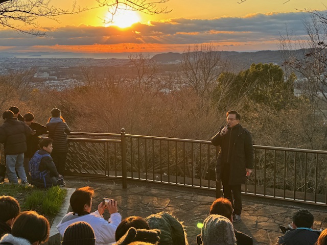 初日の出をバックにマイクを持って挨拶する市長。