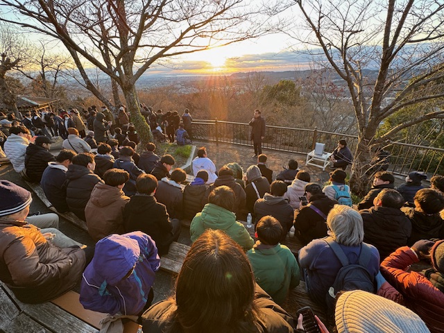 初日の出をバックに挨拶する市長と、それを見る多数の見学者。