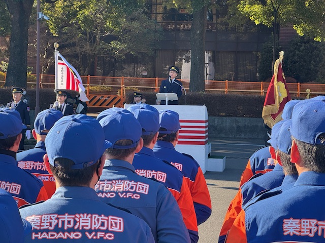 屋外で挨拶をする制服に制帽姿の市長。手前には消防団の制服を着た団員が多数整列している。