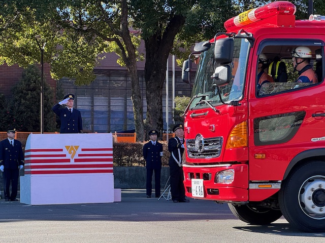 白地に黄色の市章が入った演台に立って敬礼する市長。手前を消防車が通っている。