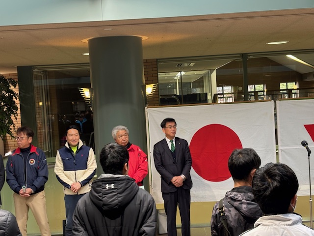 日の丸の旗の前で立つスーツの市長と、その左側にジャージやウィンドブレーカーを着た来賓の男性3人