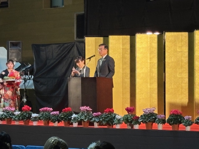 ステージの上で挨拶する市長。ステージの端にはシクラメンが隙間なく並び、背後には金屏風が置かれている。