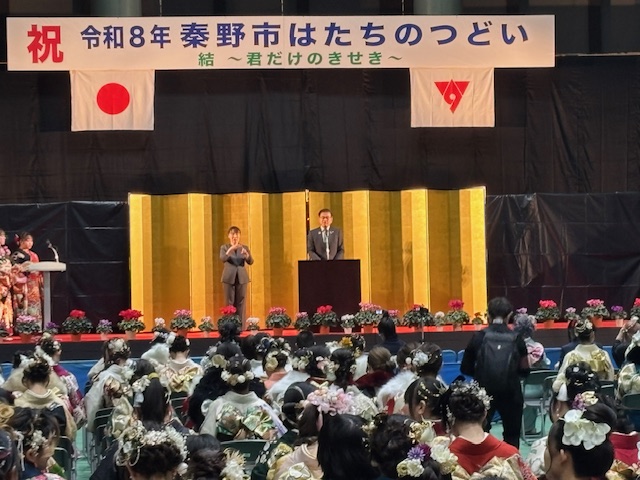 金屏風を背に挨拶する市長。左隣には手話通訳者が立っている。左端には司会をする振り袖姿の女性。ステージ下には多数の振り袖姿の出席者。