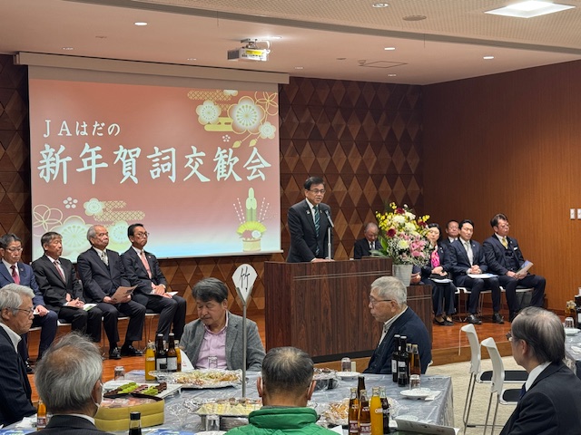 賀詞交歓会の文字が写ったスクリーンの前で挨拶をする市長。左側に主催者の男4名、右側に来賓が8名ほど座る。